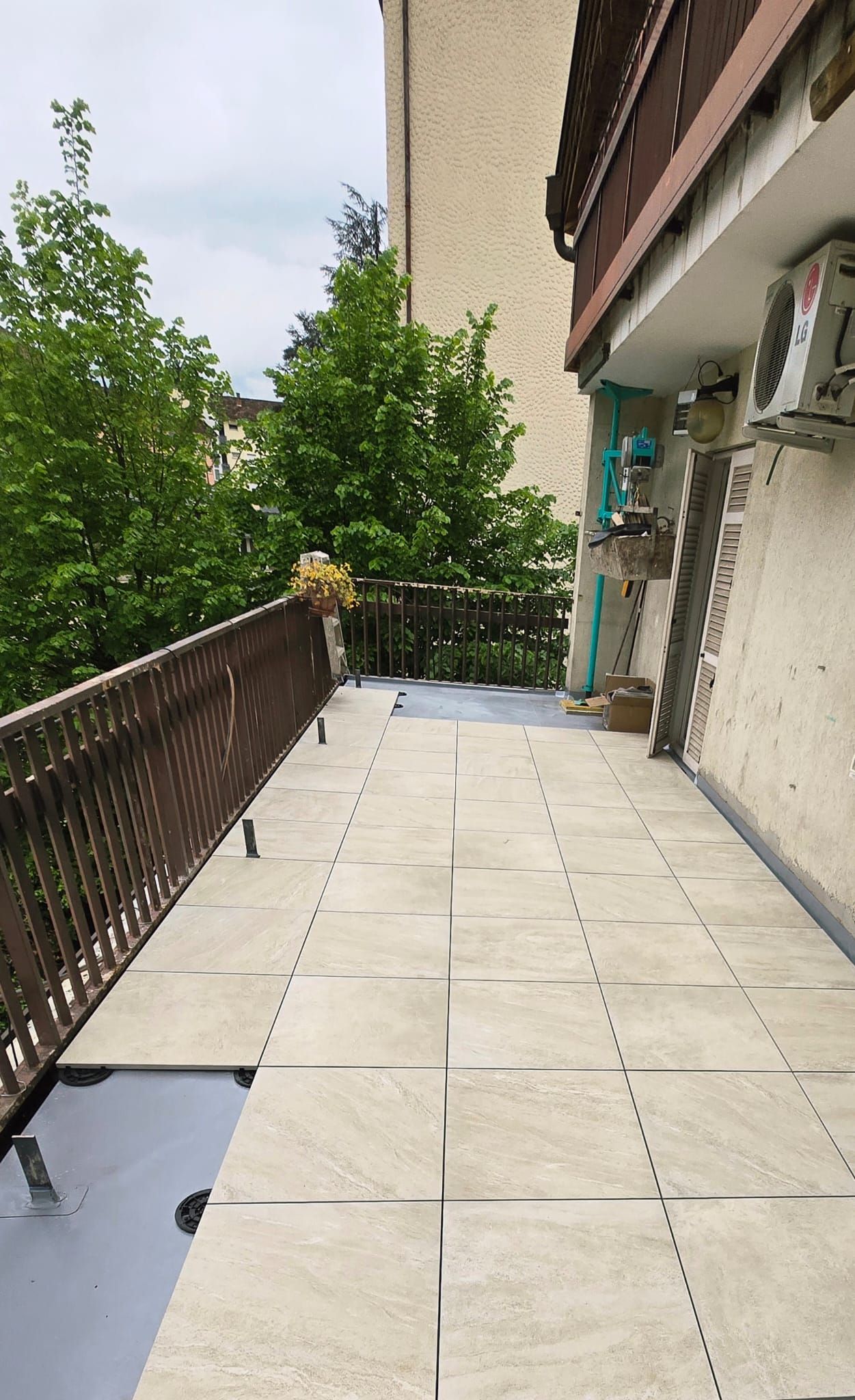 Balcone con pavimento in pietra, ringhiera in metallo e vista sugli alberi. Edifici ai lati.