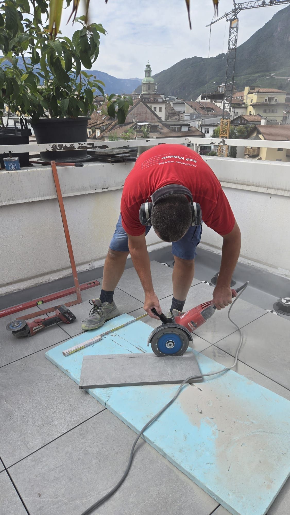 Persona con una maglietta rossa che usa una sega elettrica per tagliare una tegola su un tetto con le montagne sullo sfondo.