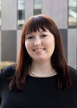 A woman in a black dress and pearl necklace is smiling in front of a building.