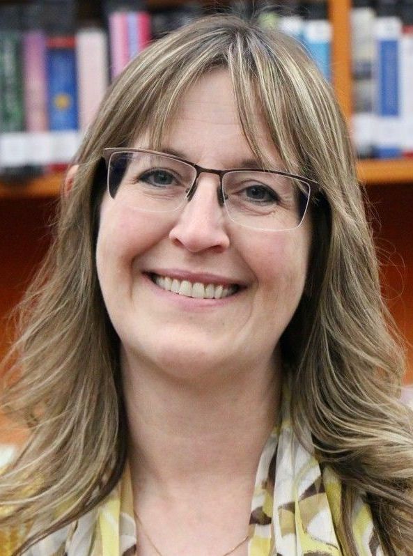 A woman wearing glasses is smiling in front of a bookshelf