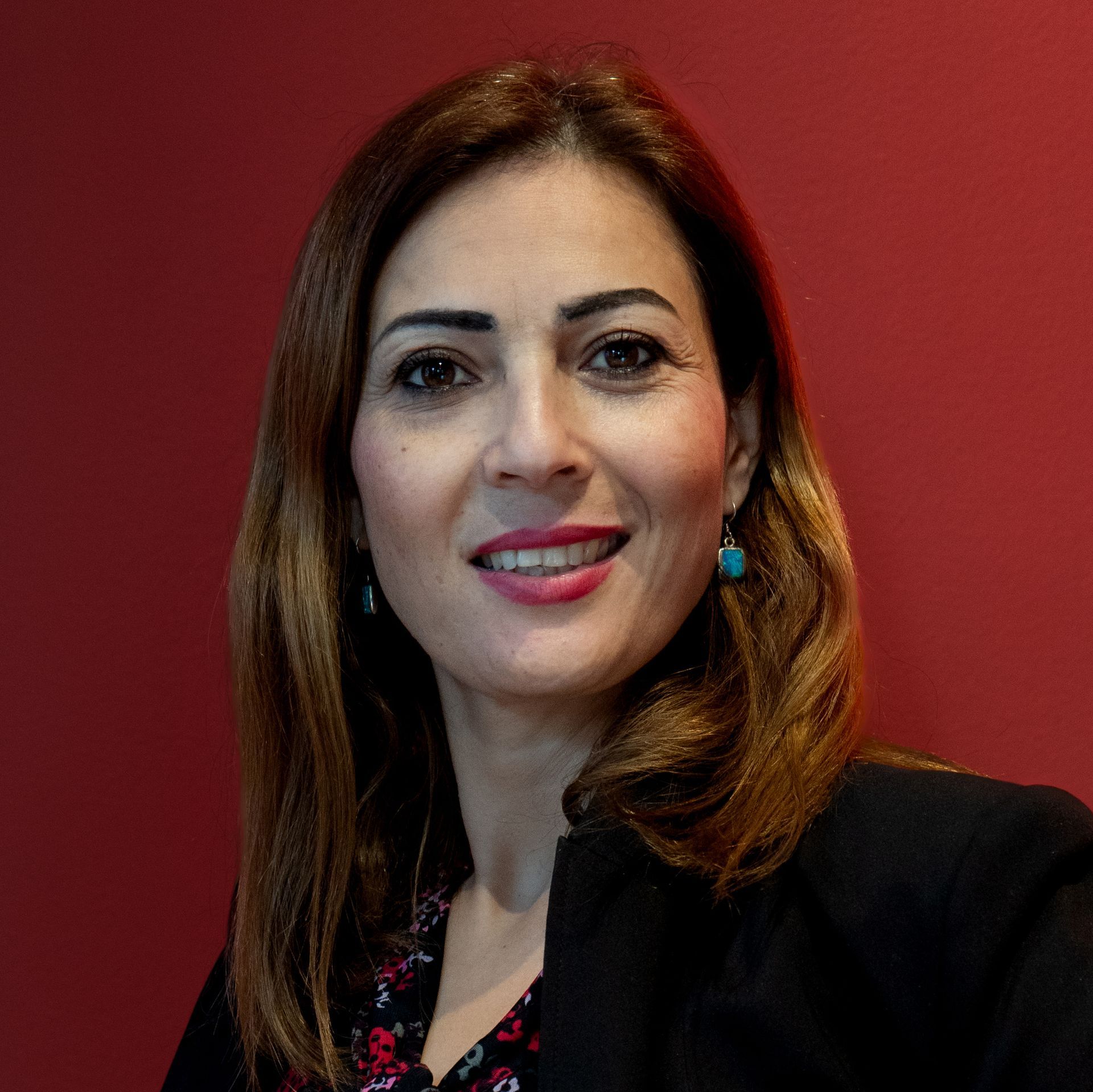 A woman wearing a black jacket and earrings is smiling in front of a red wall.