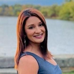 A woman in a blue tank top is smiling in front of a body of water.