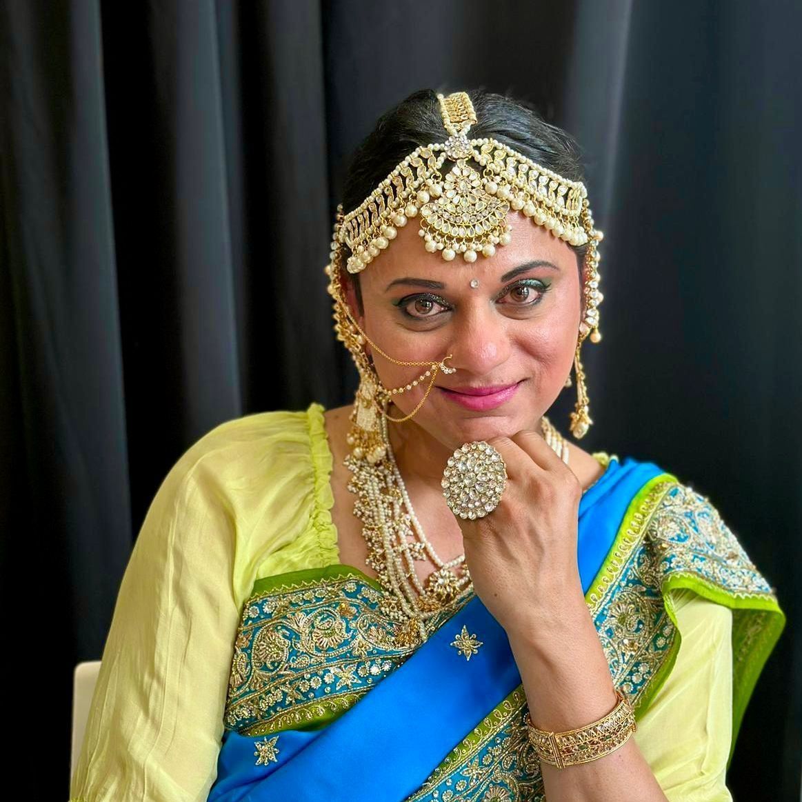 A woman wearing a blue and yellow saree and gold jewelry is sitting in front of a black curtain.