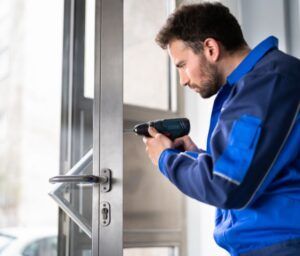 A man in a blue jacket is using a drill to fix a door.