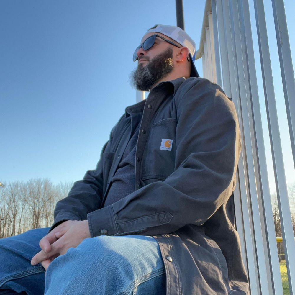 A man with a beard is sitting on a railing wearing sunglasses and a hat