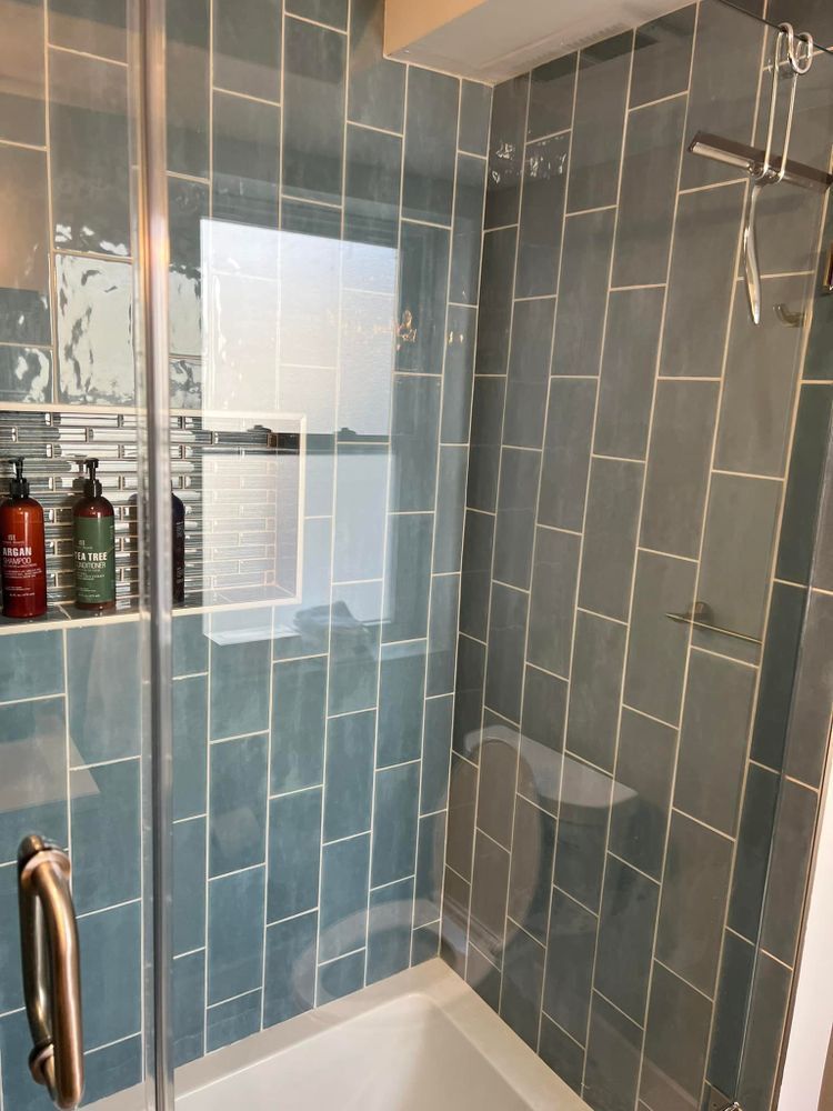 A bathroom with blue tiles , a tub , a shower and a window.