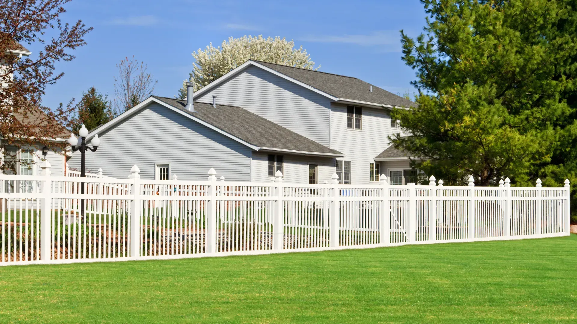 White picket fence surrounds a two-story gray house with a green lawn and trees on a sunny day.