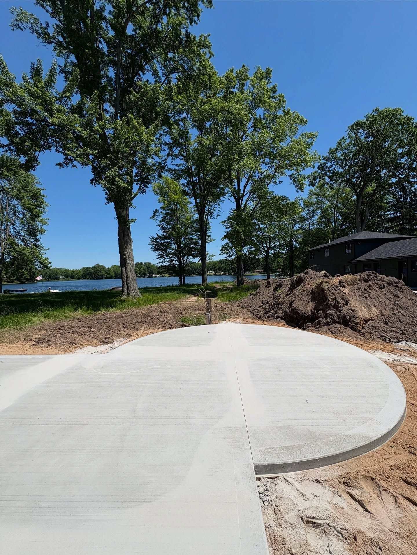Concrete path under construction by a lakeside, with trees, dirt piles, and a blue sky