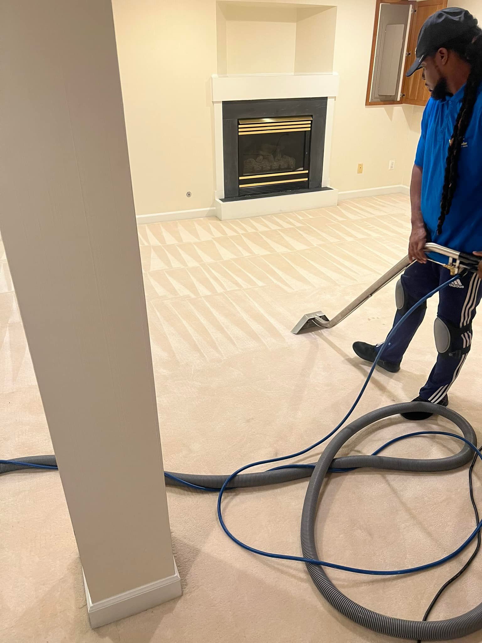 A person in blue outfit steam cleans a beige carpeted room with fireplace.