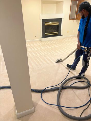 A person in blue outfit steam cleans a beige carpeted room with fireplace.