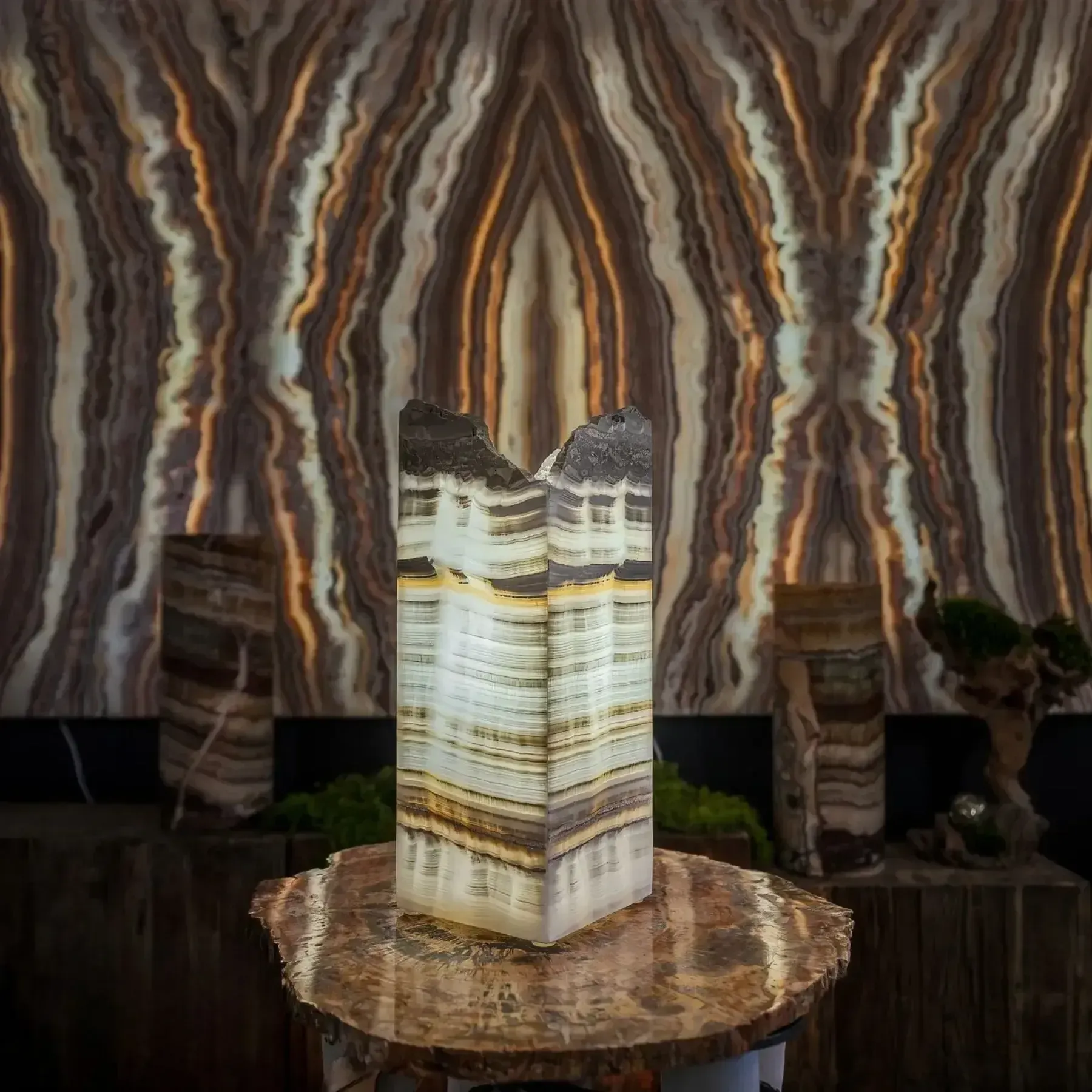A backlit, vertically banded stone sculpture sits on a wooden pedestal, set against a large, matching stone wall backdrop.