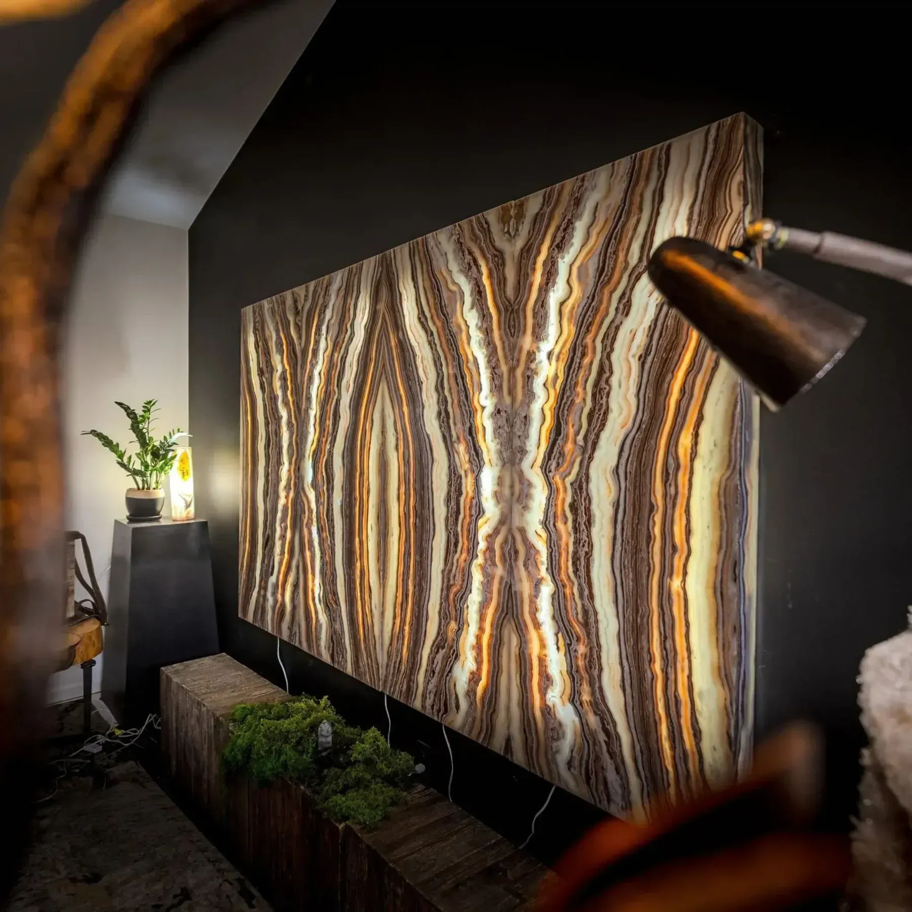 Backlit, bookmatched onyx wall panel with brown and beige veining, set on a dark wall with ambient room lighting.