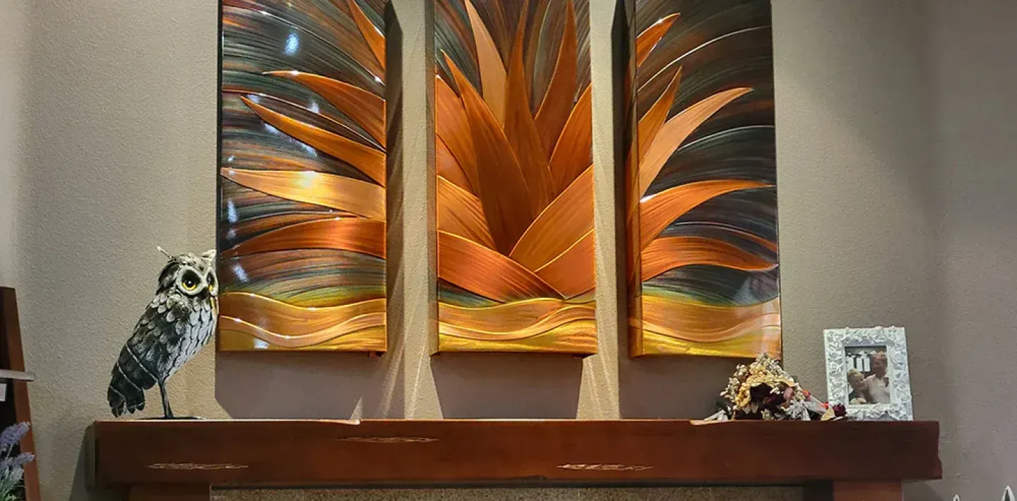 Three abstract orange and bronze agave art panels hanging above a wooden fireplace mantle with an owl statue and frame.