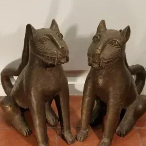 Two bronze-colored, stylized feline sculptures sit side-by-side on a red tiled floor against a white wall.