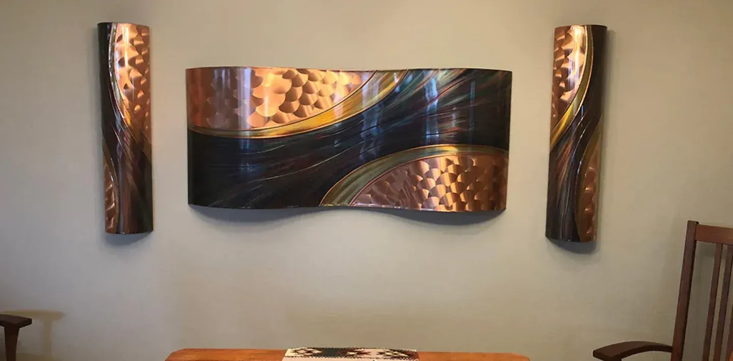 Three pieces of abstract metal wall art with copper and dark swirls mounted on a grey wall above a wooden table.