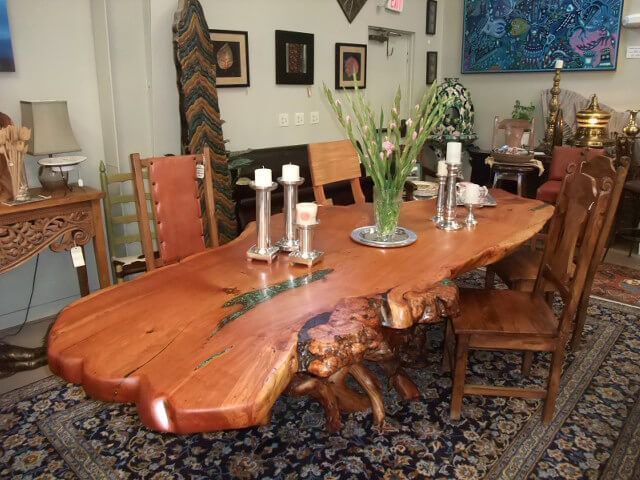 A long, reddish-brown live-edge wooden dining table with a natural root base, surrounded by chairs on a patterned rug.
