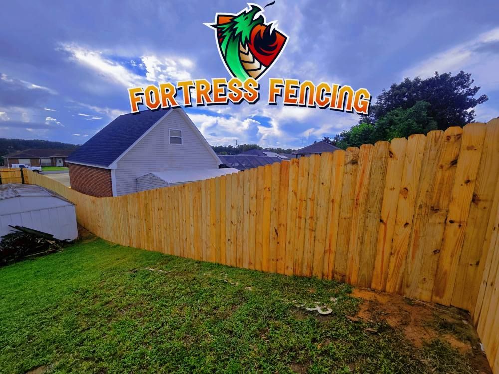 Wooden fence surrounding a yard with a house and cloudy sky in the background. The Fortress Fencing logo is at the top.