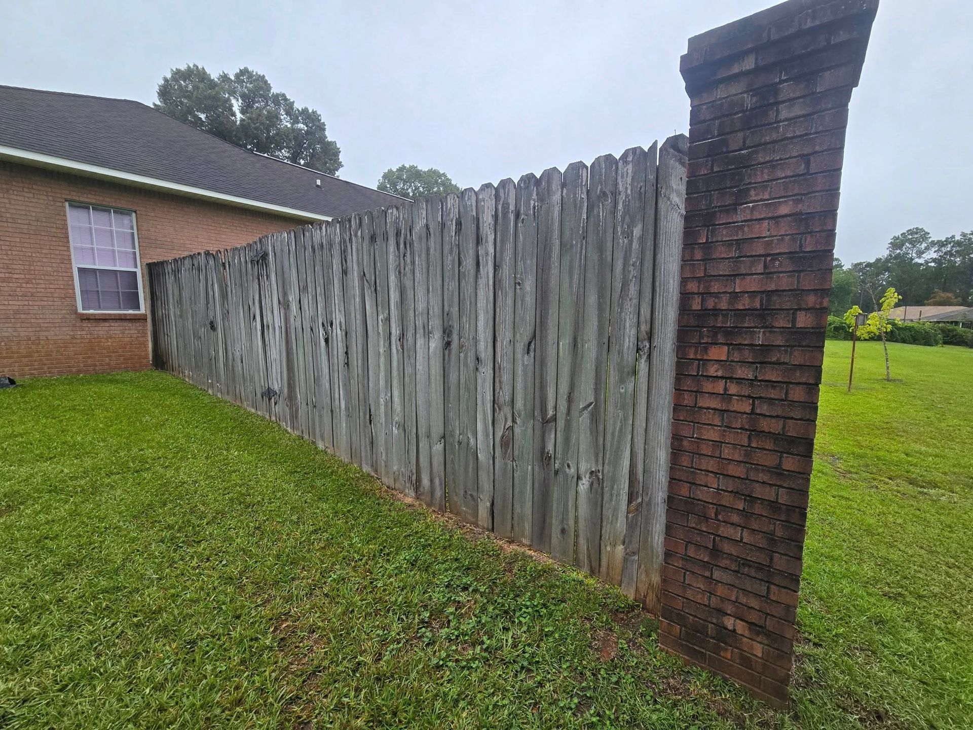 Wooden fence next to a brick pillar, near a house and grass lawn.
