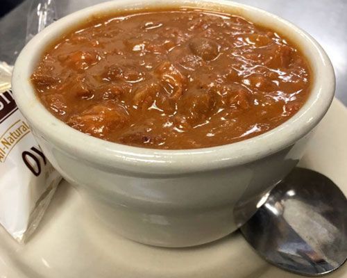 Bowl of chili on a saucer with a spoon and a packet of crackers, all on a diner table.