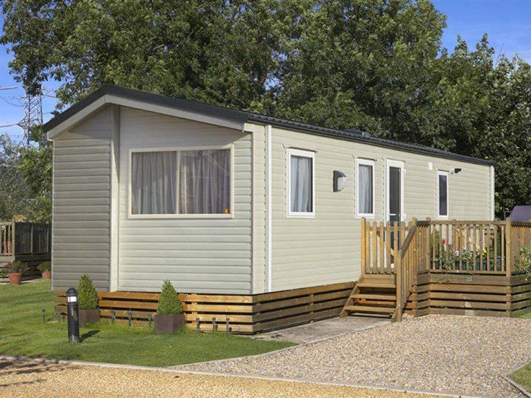 A mobile home with a wooden deck and stairs is parked in a gravel lot.