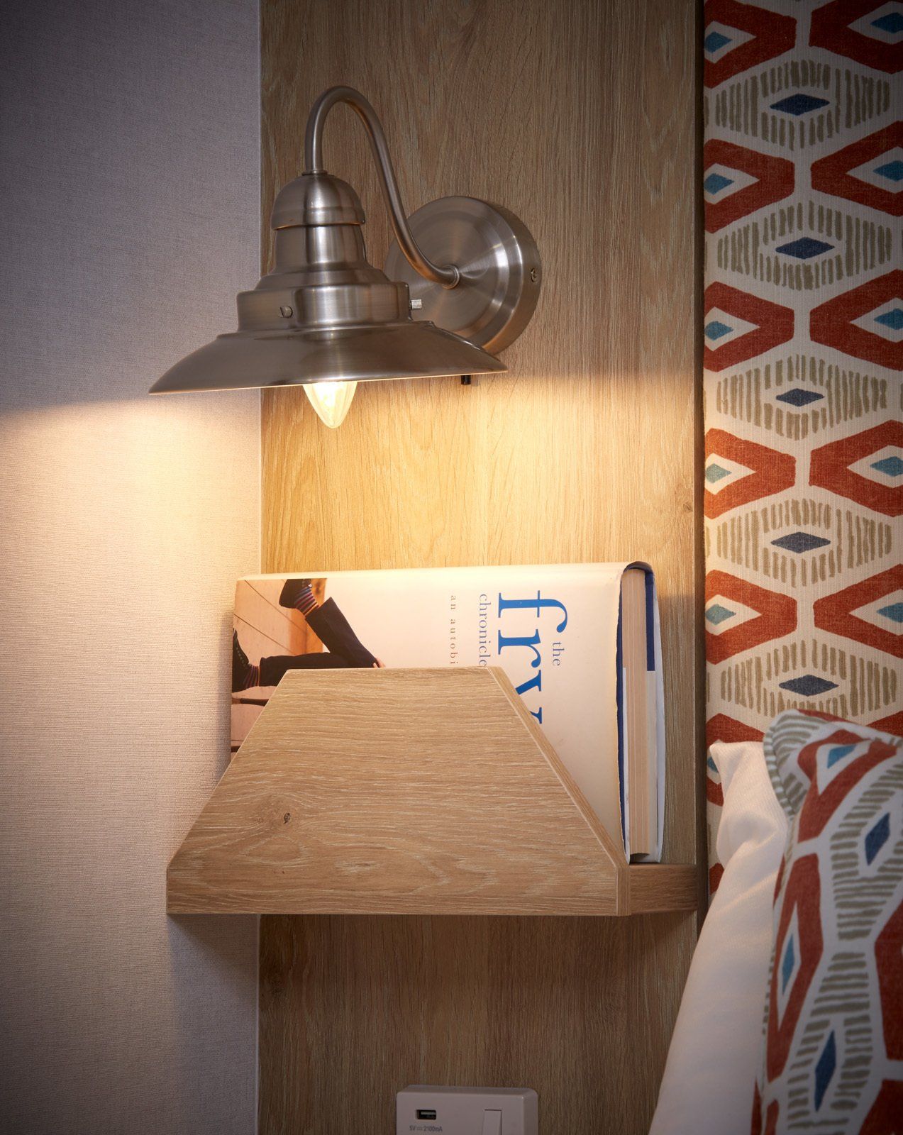 A book is sitting on a wooden shelf next to a lamp.