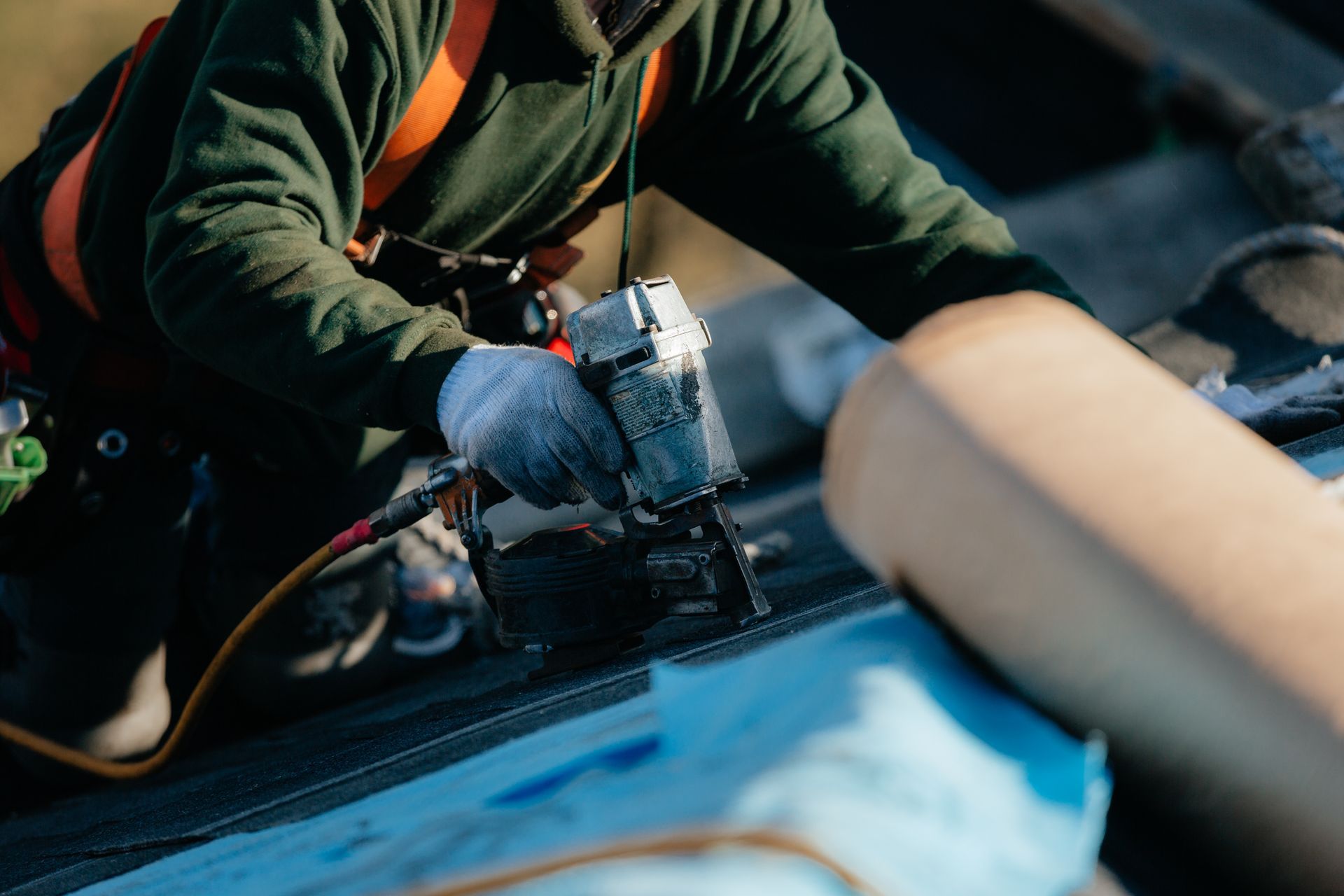 Couvreur en veste verte, installant des matériaux de toiture avec un pistolet à clous tout en portant un harnais de sécurité.