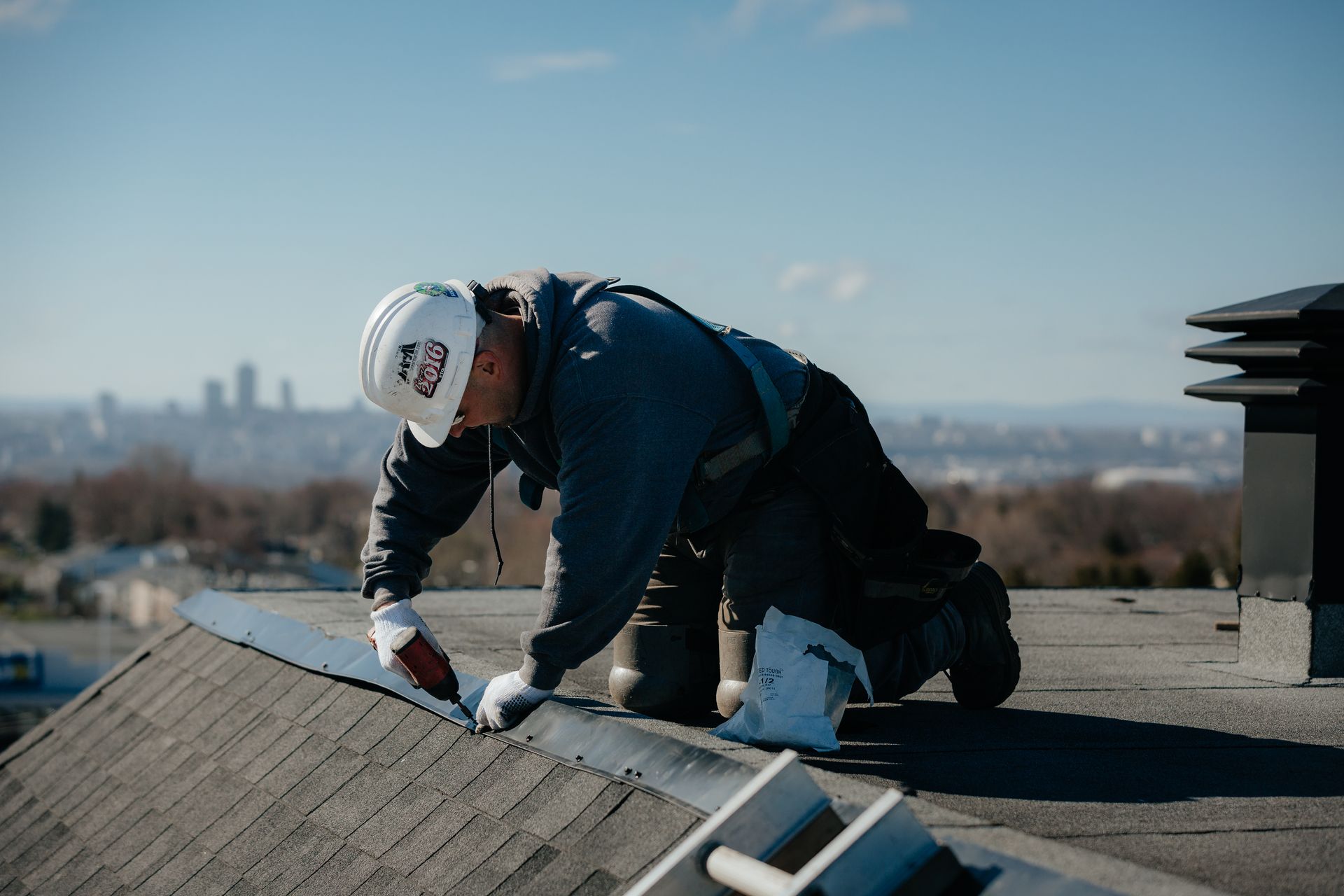 Couvreur sur un toit, appliquant du mastic. Il porte un casque et des gants, avec la silhouette de la ville en arrière-plan.