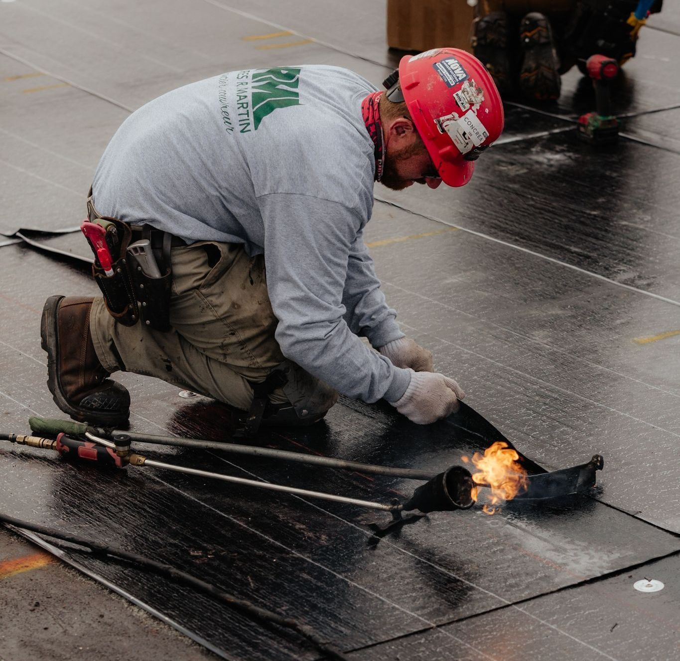 Un couvreur portant un casque de sécurité soude des matériaux de toiture avec un chalumeau à l'extérieur.