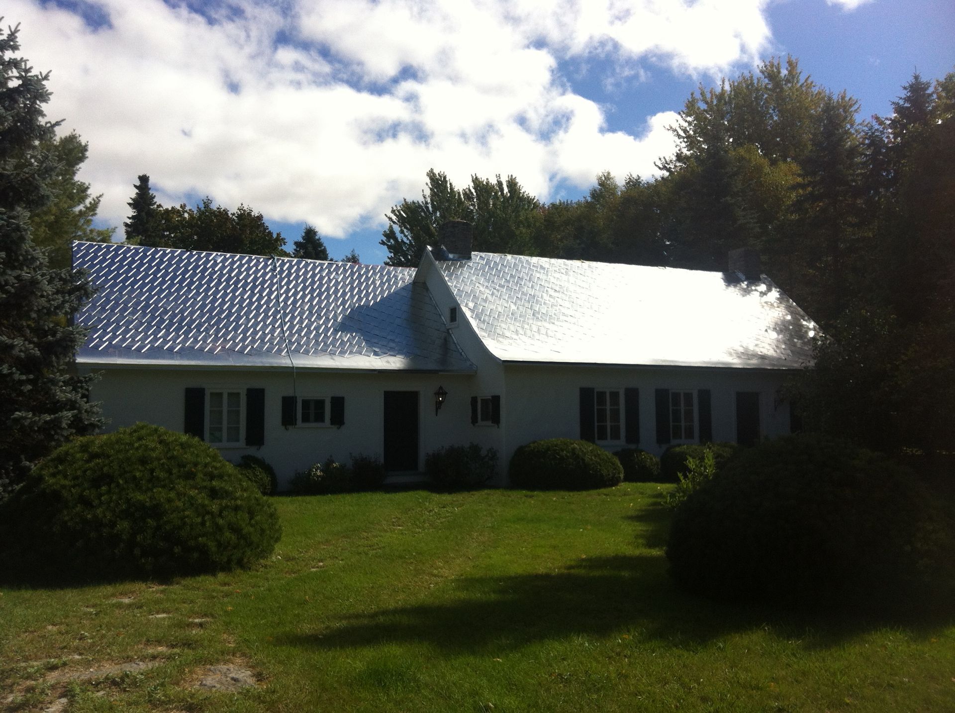 Maison blanche avec un toit argenté, des buissons verts et un ciel bleu.