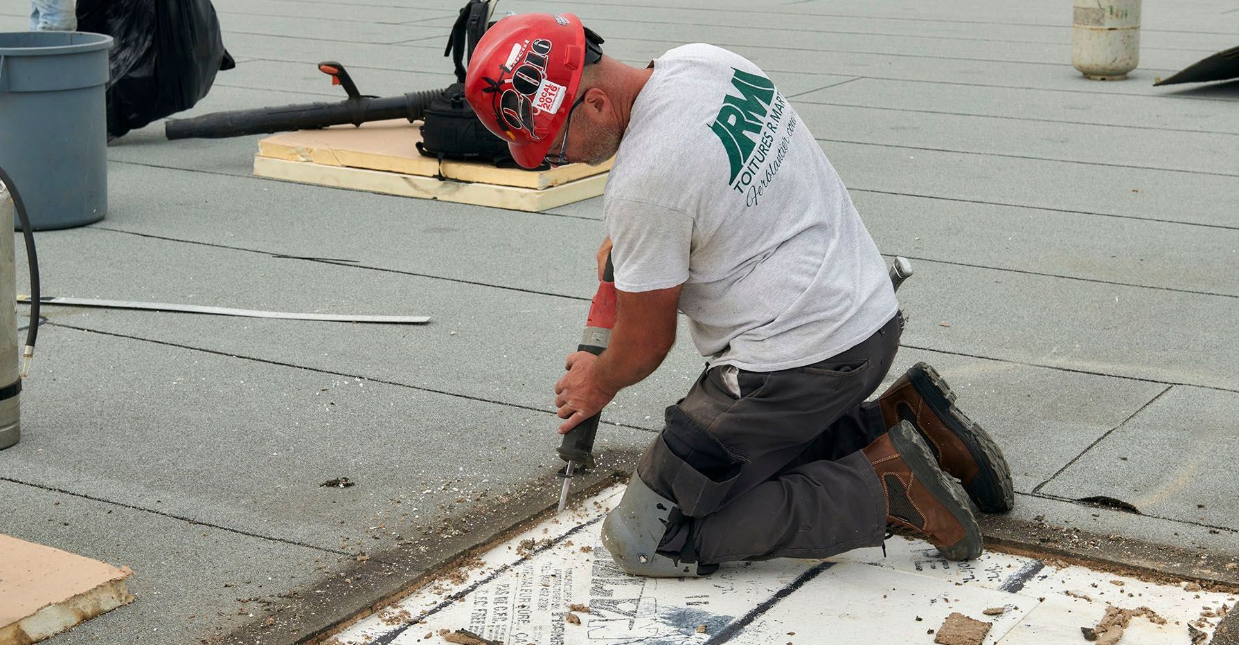 Ouvrier du bâtiment sur un toit plat, à genoux et utilisant une perceuse ; tons gris, blanc et marron.