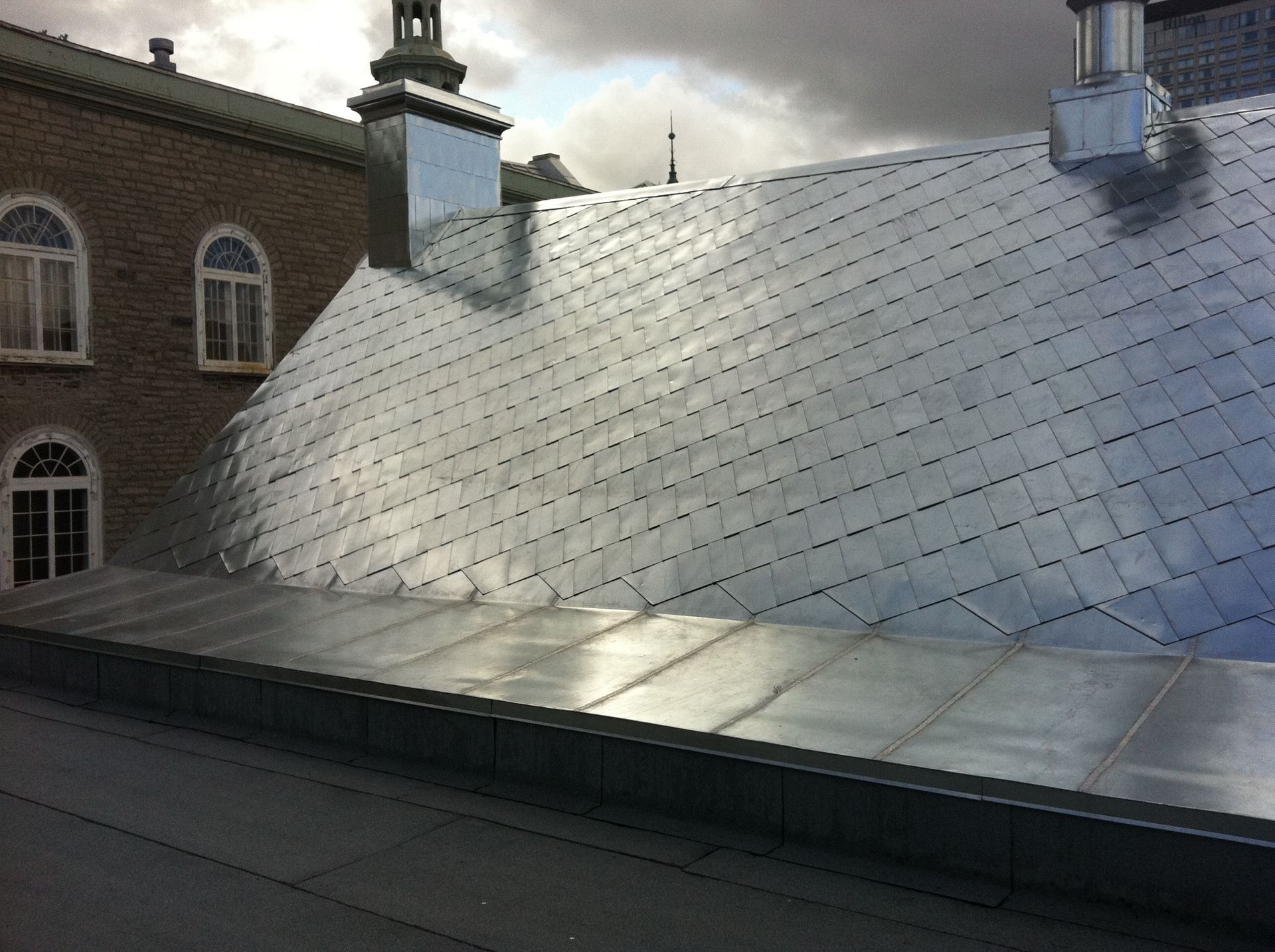 Toit en métal brillant sur un bâtiment avec des fenêtres cintrées et un ciel nuageux.