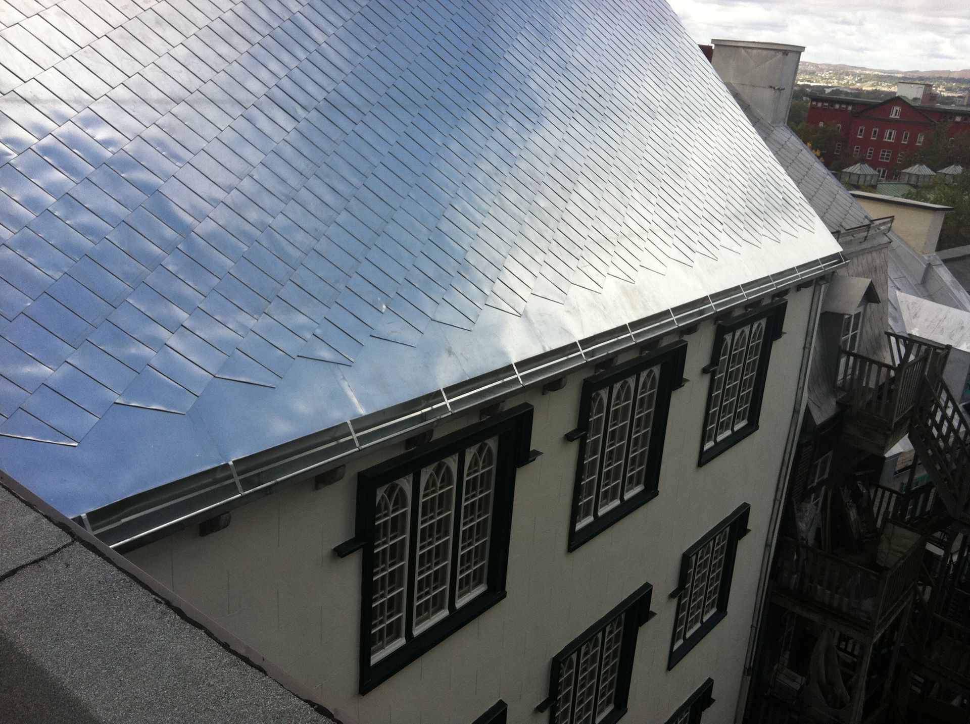Bâtiment avec toit métallique réfléchissant, fenêtres à cadre sombre et gouttière ; ciel reflété dans le toit.