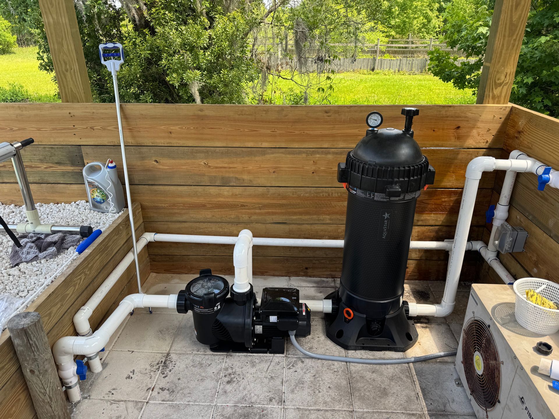 Pool equipment setup within a wooden enclosure; includes a pump, filter, pipes, and gauge, outdoors.