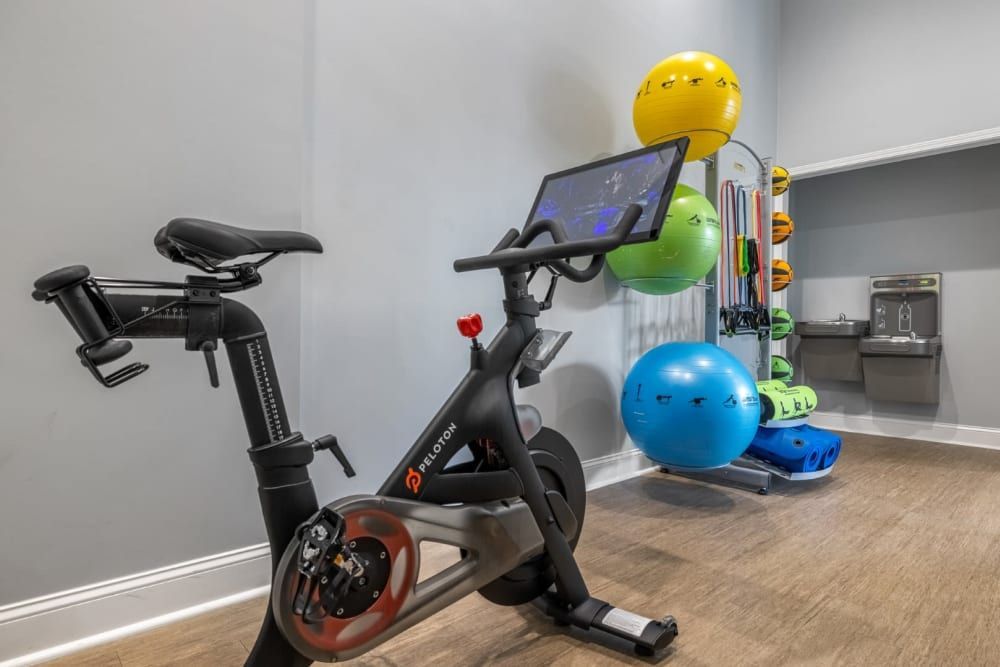 Image of a modern indoor gym featuring a stationary cycling bike in the foreground with a digital screen. In the background, there is a rack holding various colored exercise balls and resistance bands. A water cooler is visible towards the right. The room has wooden flooring and light gray walls at Marq on Ponce.