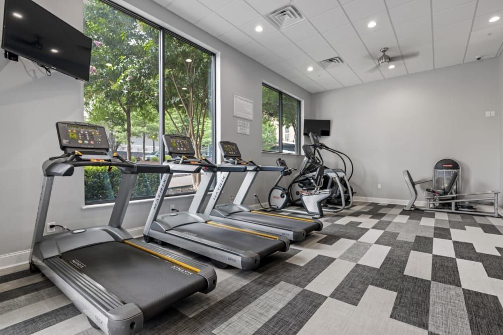A well-equipped modern gym interior with three treadmills in the foreground, an elliptical trainer, and an exercise bike towards the back. The fitness room features large windows with a view of trees, a patterned black-and-white checkered floor, and a television mounted on the wall to the left, offering a comfortable and inviting space for exercise and fitness activities at Marq on Ponce.