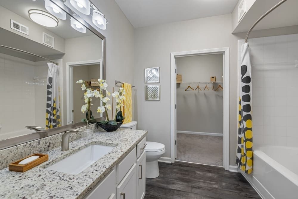 A modern bathroom interior featuring a large vanity with granite countertop and two sinks under a lit wall-mounted light fixture. A decorative orchid, a bar of soap in a wooden tray, and an oval mirror enhance the space. A shower curtain with a yellow and gray pattern partially conceals a bathtub to the right. Beyond the open door, a glimpse of a neat walk-in closet with wooden shelves and hangers is visible. The floor is styled with dark wood-look flooring, and the walls are painted in a light neutral tone at Marq on Ponce.