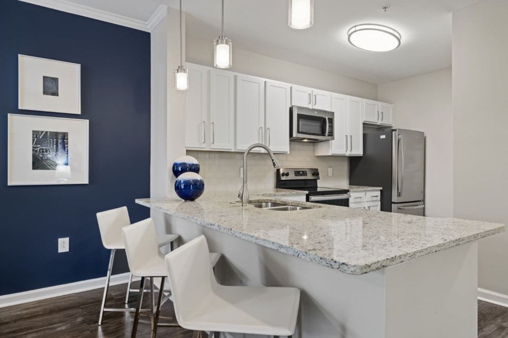 Modern kitchen interior with white cabinetry and stainless steel appliances. Features a central island with a granite countertop and white bar stools, pendant lighting, and a navy blue accent wall with framed artwork at Marq on Ponce.