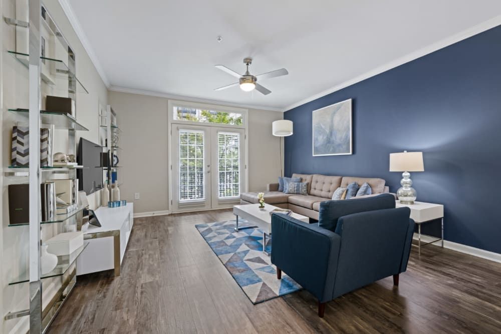 Modern living room with a white and navy blue color scheme, featuring a tufted beige sofa with blue throw pillows, a navy blue armchair, and a white coffee table on a geometric patterned blue area rug. The room is accented with a dark blue accent wall, upon which a large abstract painting is hung, a glass shelving unit filled with decorative items, and white French doors with white curtains leading to an outdoor area. The space is well-lit with natural light coming through the doors and a ceiling fan with a light fixture above at Marq on Ponce.