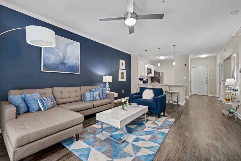 Modern living room with a beige sectional sofa accented with blue pillows, a white coffee table on a vibrant blue and white area rug, and a dark blue accent wall featuring a large abstract painting. The space includes a white lamp with an arched stand, a ceiling fan, and transitions into a kitchen with bar seating and stainless steel appliances. The overall color scheme is neutral with blue decor accents, creating a contemporary and welcoming atmosphere at Marq on Ponce.