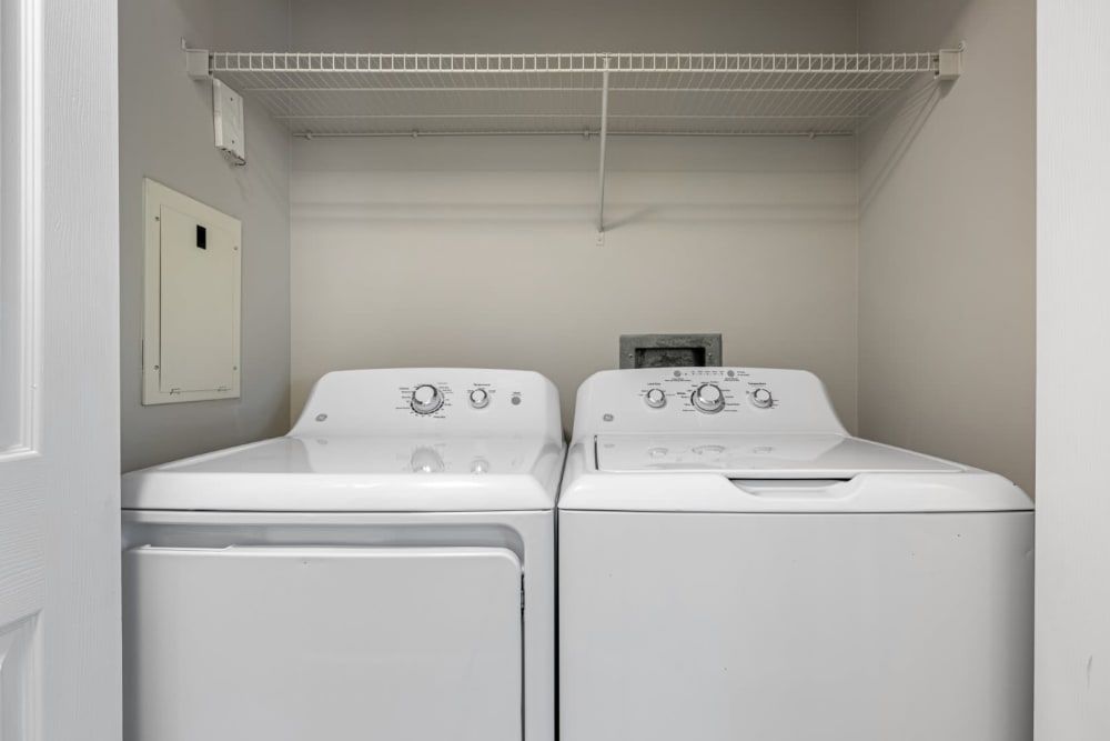 Laundry room interior with a white top-loading washing machine on the left and a white front-loading dryer on the right, both under a wire shelf. A small closed utility or fuse box is visible on the wall to the left at Marq on Ponce.