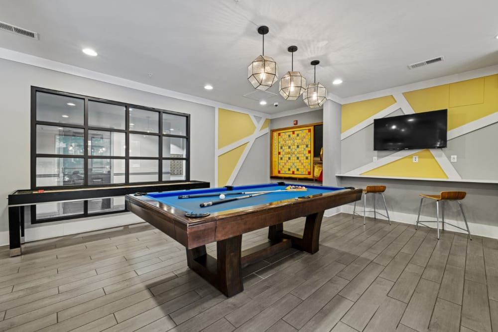 A modern game room interior with a wooden pool table with blue cloth in the foreground, a black foosball table by a large window, geometric yellow and gray accent walls, mounted flatscreen TV, and pendant lighting fixtures above. To the right, there's a high bar table with stools at Marq on Ponce.