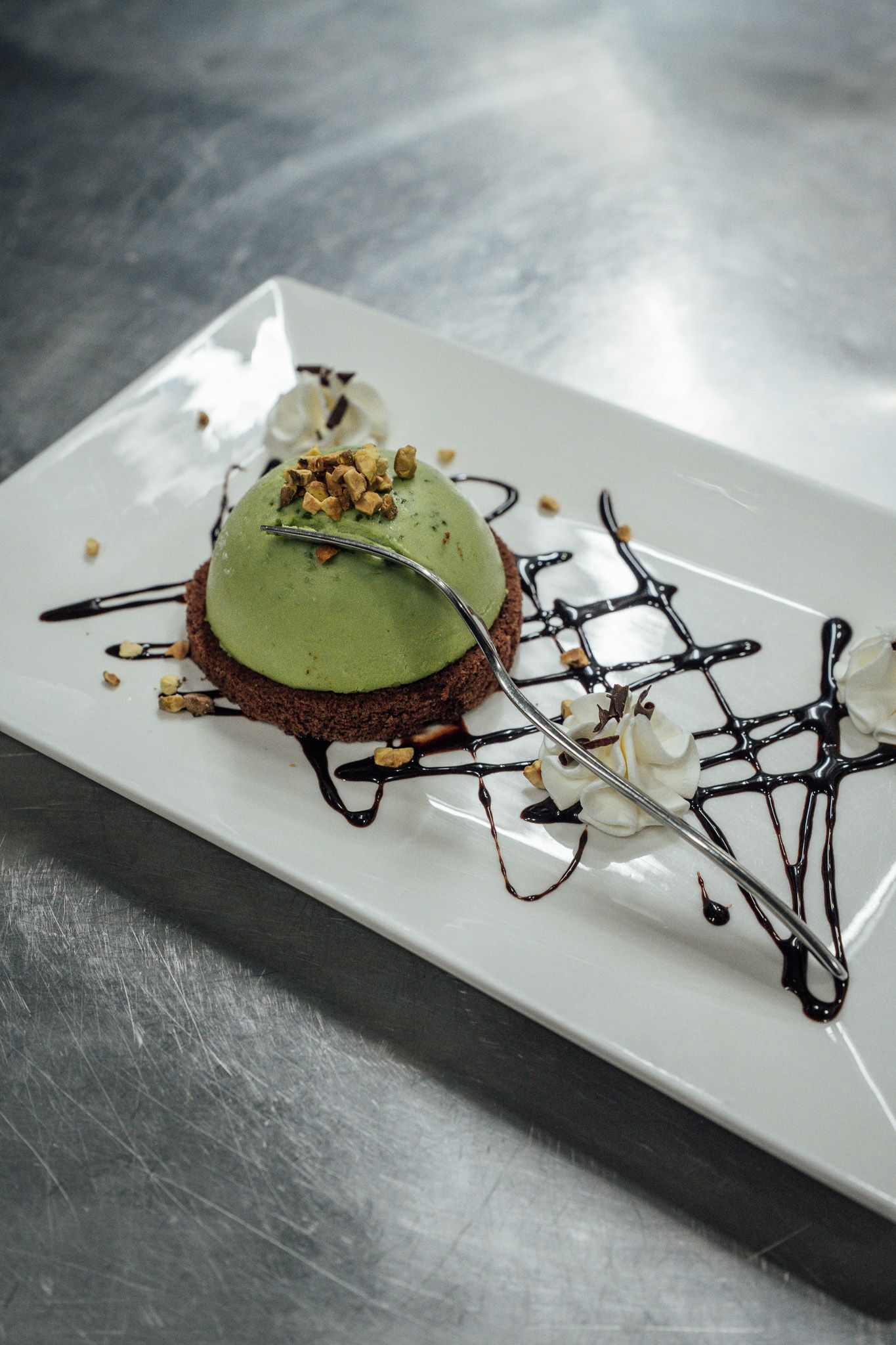 Una cupola verde per dessert su una base marrone scuro, ricoperta di cioccolato e guarnita con noci e panna montata, su un piatto bianco.