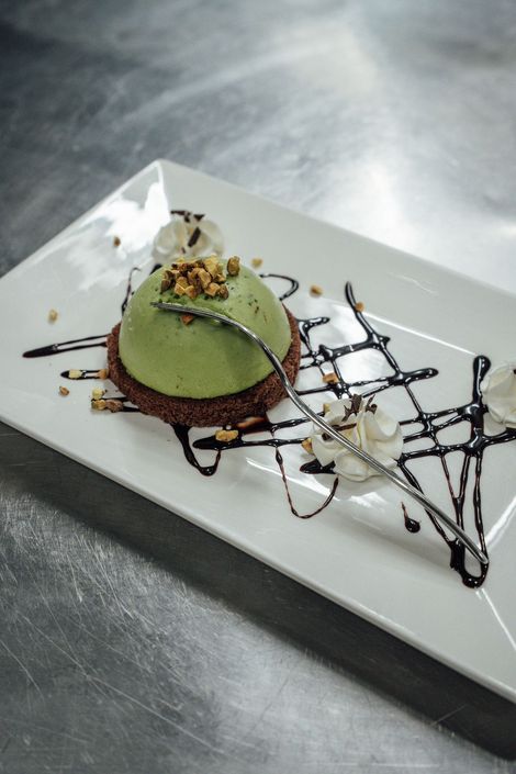 Una cupola verde per dessert su una base marrone scuro, ricoperta di cioccolato e guarnita con noci e panna montata, su un piatto bianco.