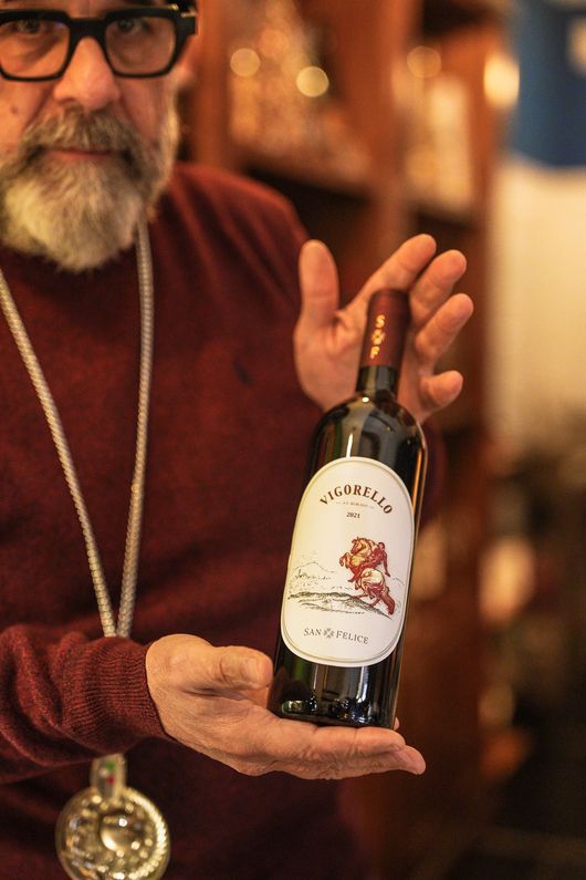 Uomo che tiene in mano una bottiglia di vino rosso; l'etichetta raffigura una scena costiera.