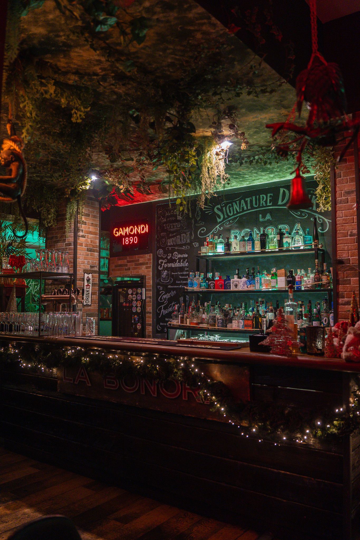 Bar buio con vegetazione lussureggiante, pareti in mattoni e luci scintillanti. Scaffali pieni di bottiglie. Insegne al neon luminose.