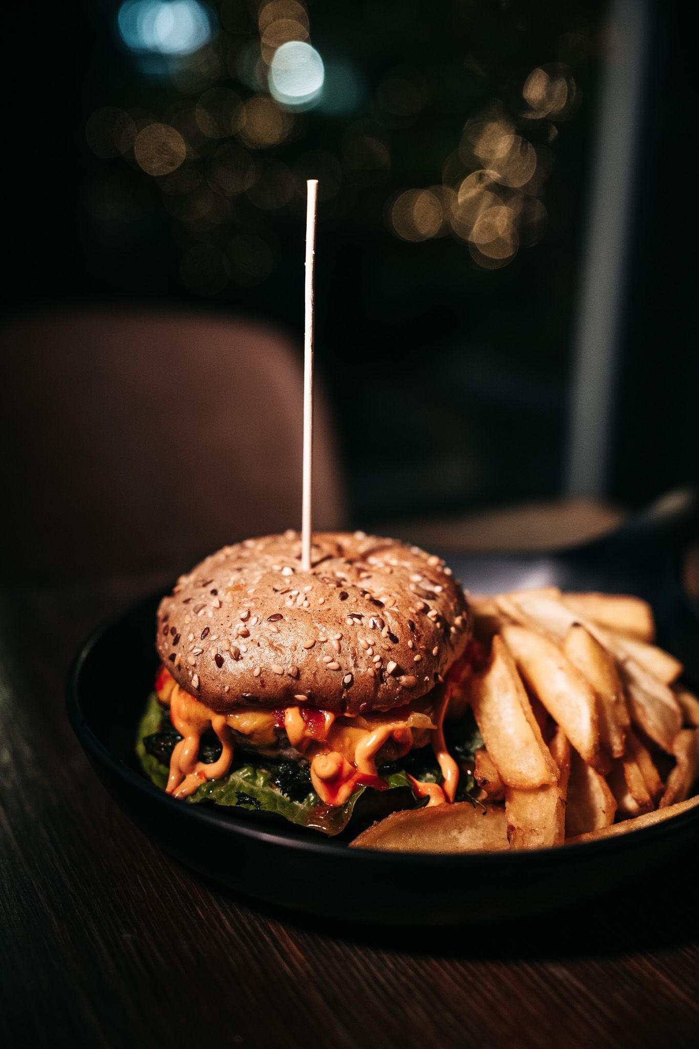 Hamburger con patatine fritte servito su un piatto nero, in una stanza buia. L'hamburger ha uno stuzzicadenti.