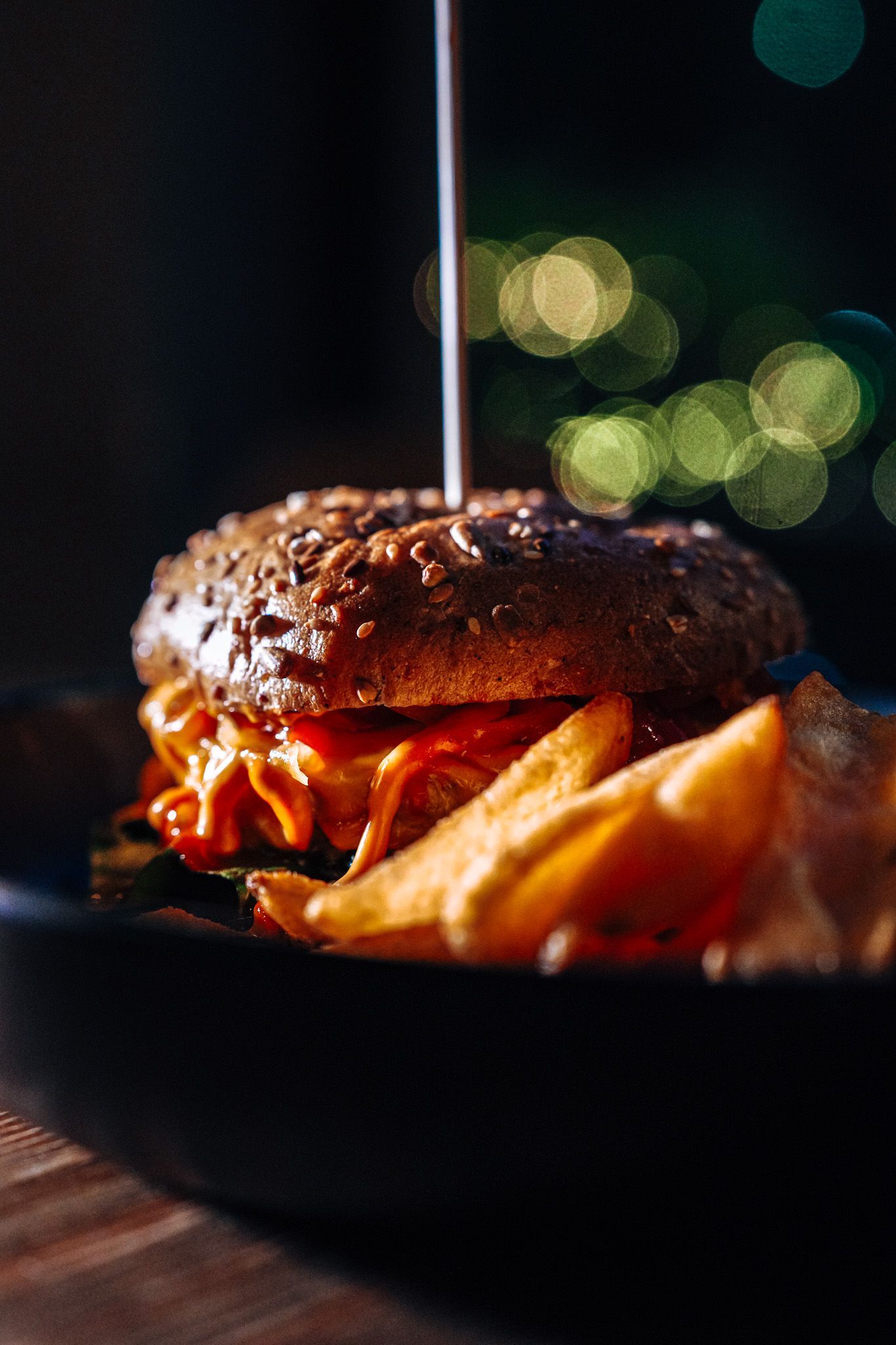 Hamburger con patatine fritte servito su un piatto scuro, illuminato da una luce soffusa. Attenzione al panino con semi di sesamo e agli spicchi di patate.