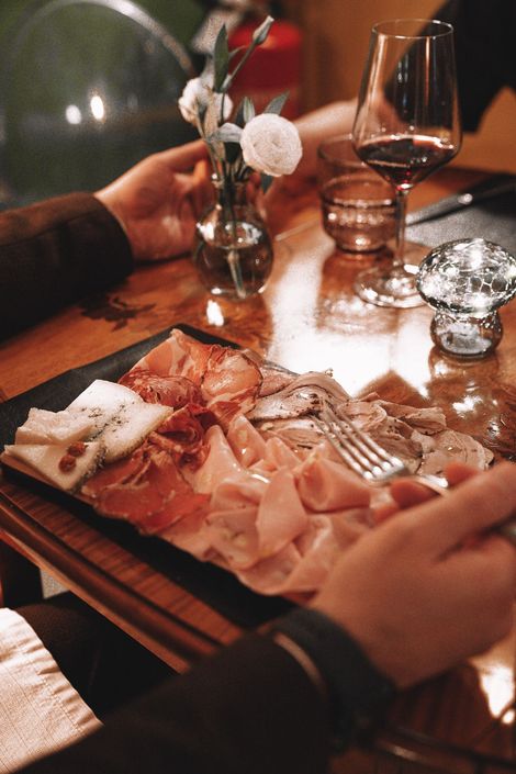 Piatto di salumi su un tavolo di legno, persona che allunga la mano verso il cibo, bicchiere di vino e piccolo vaso di fiori.