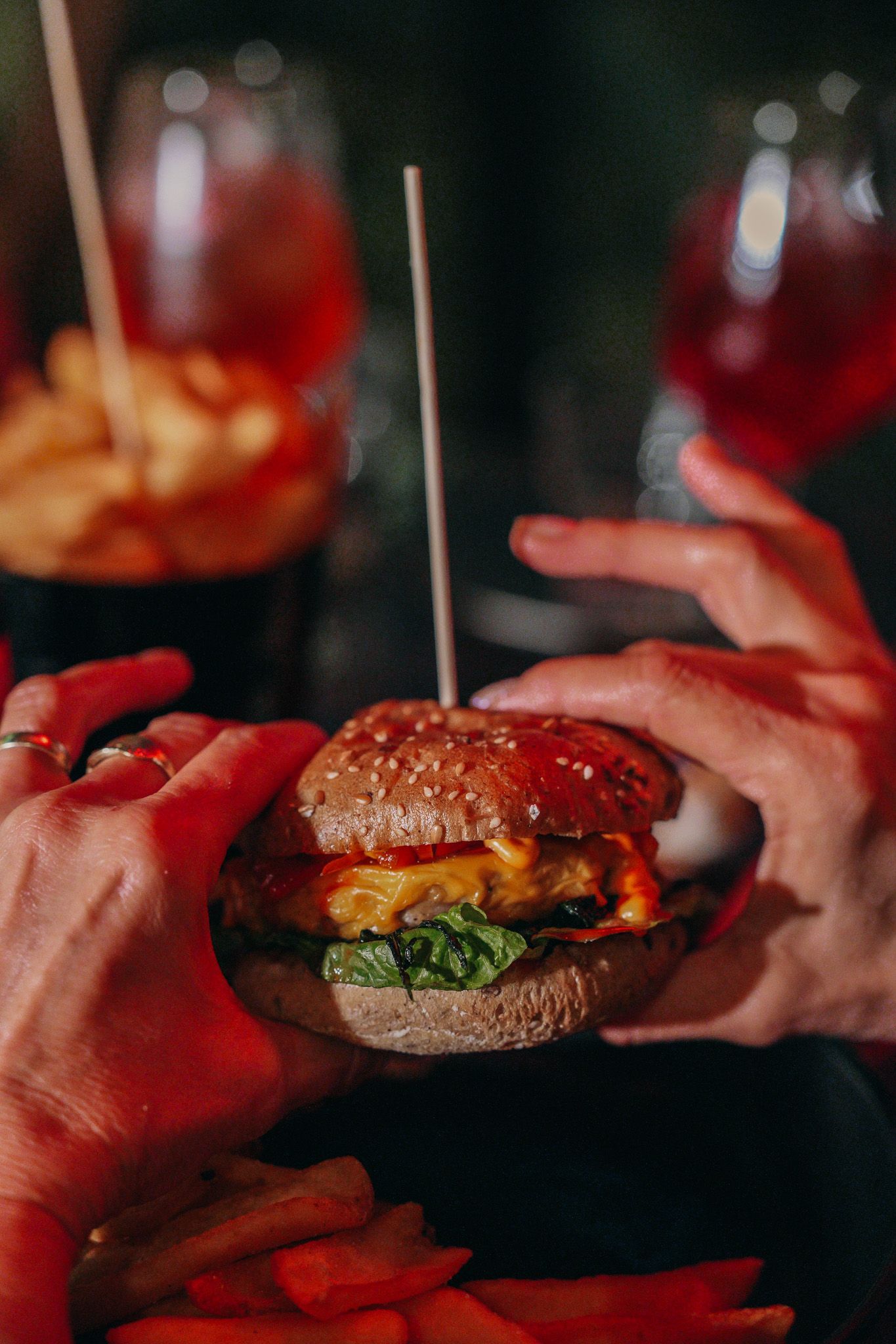 Mani che tengono un hamburger con uno stuzzicadenti; sullo sfondo patatine fritte e bevande.