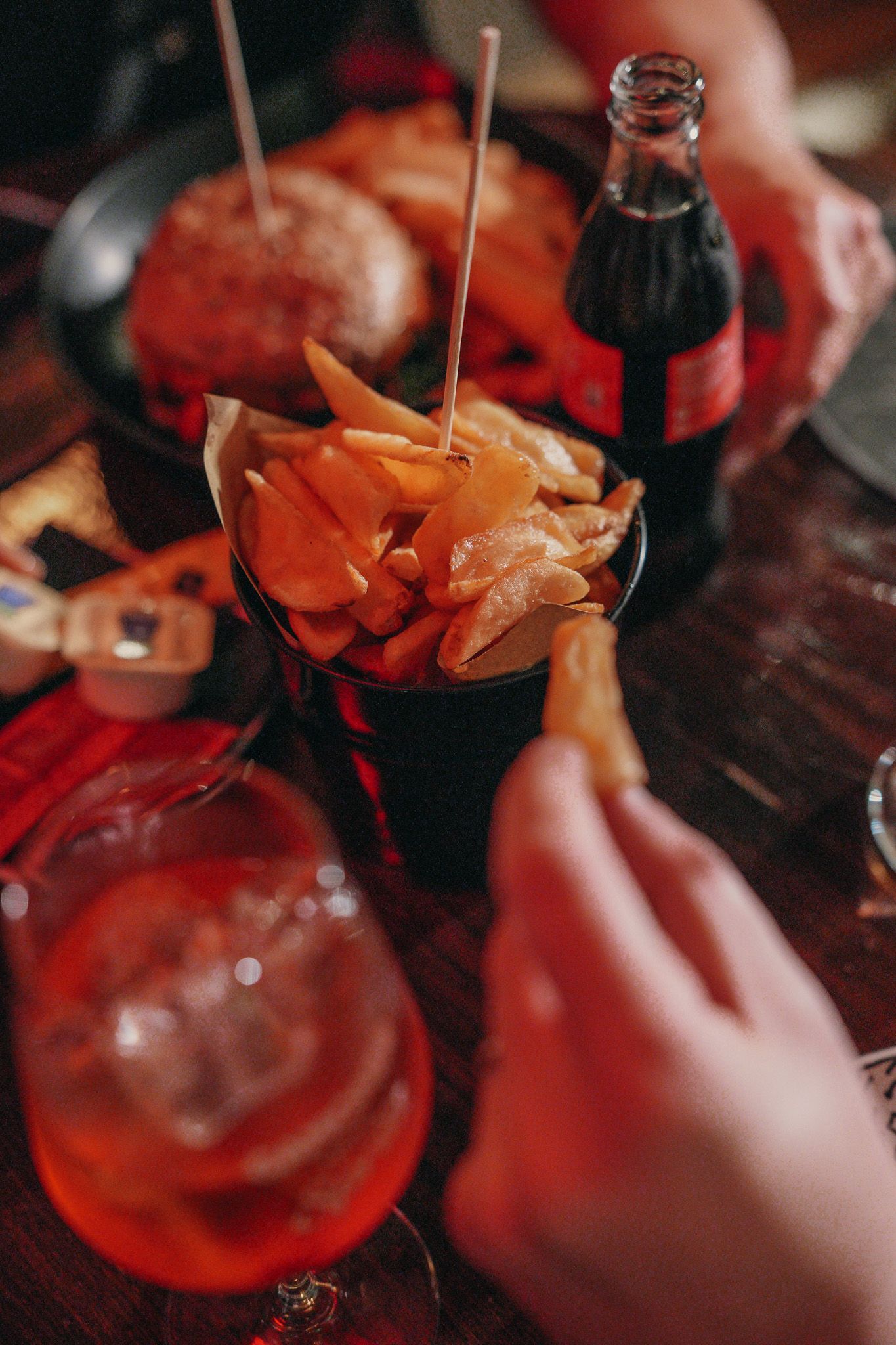 Patatine fritte in una ciotola nera, hamburger, bottiglia di soda e cocktail su un tavolo di legno scuro. Mano che allunga la mano verso una patatina.